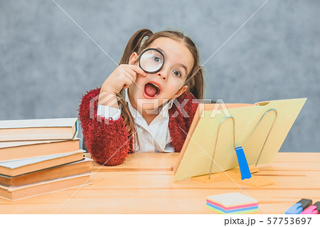 A playful little young little schoolgirl sits at the table reading a book. Her hair has two A playful little young little schoolgirl sits at the table reading a book. Her hair has two 57753697