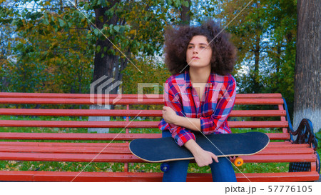 A teenage girl sits with a skateboard in her hands on a wooden park bench. 57776105