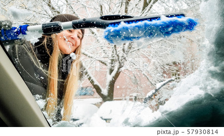 Close up shot inside the snow covered car. Young smiling woman in hat trying to clean up the Close up shot inside the snow covered car. Young smiling woman in hat trying to clean up the 57930814