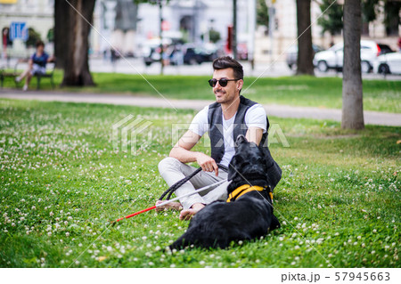 Young blind man with white cane and guide dog sitting in park in city. 57945663