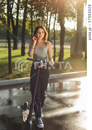 Brunette girl riding an eco-friendly electric kick scooter in a park in sunny weather on sidewalks 57993028