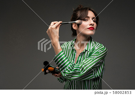 Portrait of young woman putting on makeup on cheekbones in striped blouse with makeup brushes in her Portrait of young woman putting on makeup on cheekbones in striped blouse with makeup brushes in her 58002853