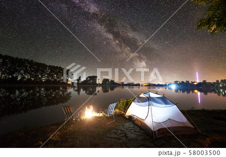 Tourist tent, two camping chairs and campfire on lake shore at night 58003500