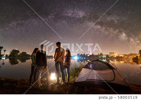 Two couple resting on lake shore near fire at night camping Two couple resting on lake shore near fire at night camping 58003519