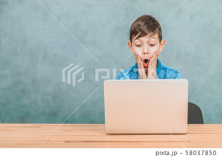 Cute little schoolboy sitting at the desk, surprised by something he has seen in his laptop. Open 58037850