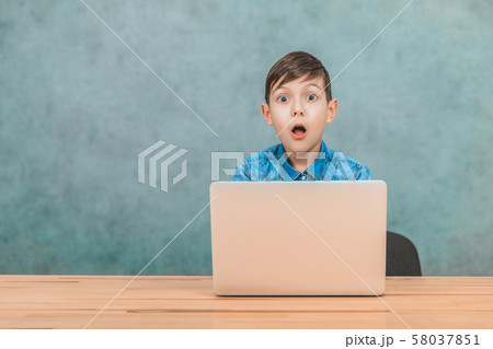 Cute little schoolboy sitting at the desk, surprised by something he has seen in his laptop. Cute little schoolboy sitting at the desk, surprised by something he has seen in his laptop. 58037851