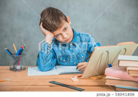 Little tired schoolboy is sitting at the desk, overwhelmed with books, leaning his head on the hand. Little tired schoolboy is sitting at the desk, overwhelmed with books, leaning his head on the hand. 58041806