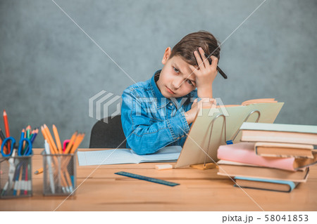 Little tired schoolboy is sitting at the desk, holding hand on the forehead, not knowing what to Little tired schoolboy is sitting at the desk, holding hand on the forehead, not knowing what to 58041853