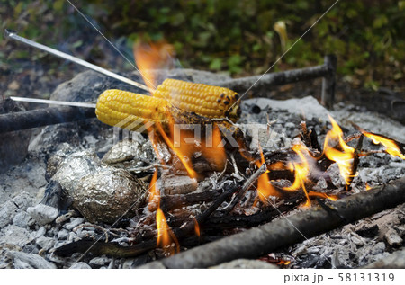 Two ears of corn roasting on the fire. The foil wrapped potatoes,  58131319