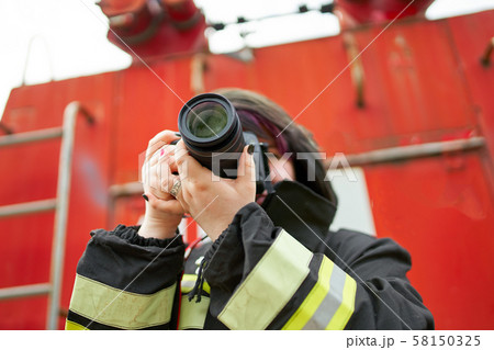 Image of young brunette fireman with photo camera in her hands on background of fire engines. 58150325
