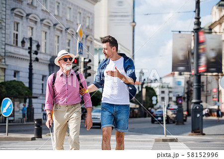 Young man and blind senior with white cane walking in city, crossing street. Young man and blind senior with white cane walking in city, crossing street. 58152996