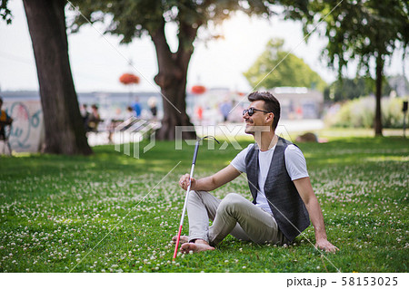 Young blind man with white cane and guide dog sitting in park in city. 58153025
