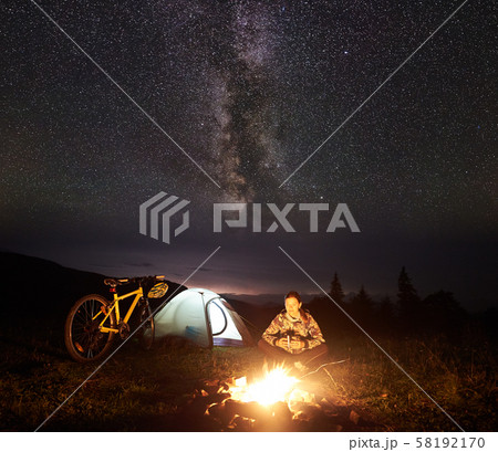 Woman resting at night camping near campfire, tourist tent, bicycle under evening sky full of stars Woman resting at night camping near campfire, tourist tent, bicycle under evening sky full of stars 58192170