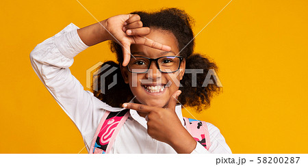 Smiling Black School Girl Looking Through Fingers Frame, Yellow Background Smiling Black School Girl Looking Through Fingers Frame, Yellow Background 58200287