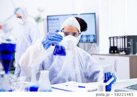 Scientist in safety glasses looks at a beaker with blue liquid and smoke 58221465