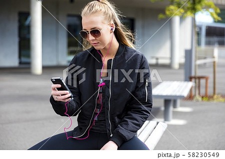Blonde woman with sunglasses sits outdoor and uses phone 58230549