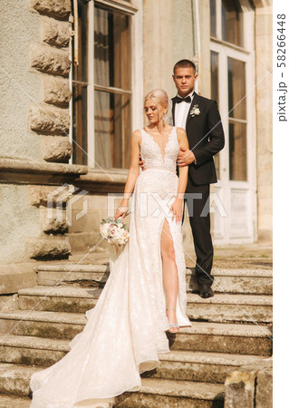 Groom and bride stand on stairs. Fashion model in elegant clothes. White long dress and stylish suit Groom and bride stand on stairs. Fashion model in elegant clothes. White long dress and stylish suit 58266448