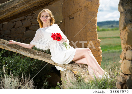 Young female in white dress posing on tree trunk with poppy flowers in hands Young female in white dress posing on tree trunk with poppy flowers in hands 58367976