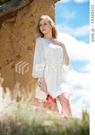 Woman wearing white dress posing near wall with poppies flowers 58368435