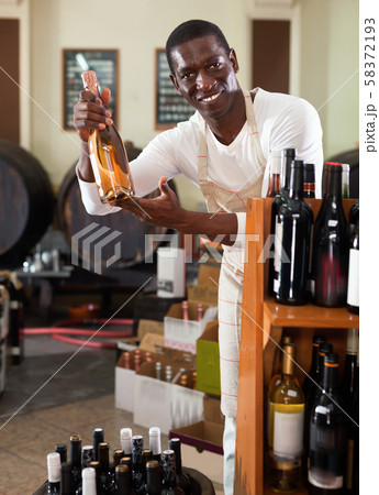Smiling salesman in apron proposing wine in bottles in winery shop Smiling salesman in apron proposing wine in bottles in winery shop 58372193