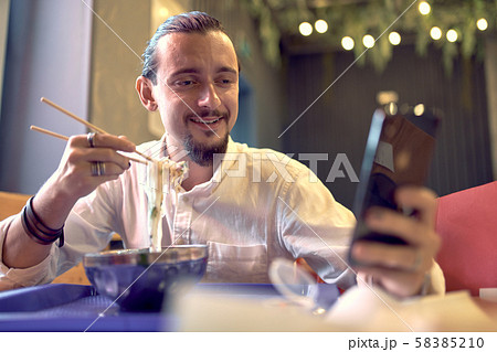 Smiling brunet man looking at phone while enjoying Asian food Smiling brunet man looking at phone while enjoying Asian food 58385210