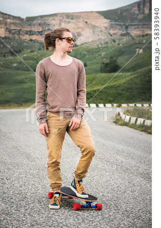 A stylish young man standing along a winding mountain road with a skate or longboard in his hands 58393940