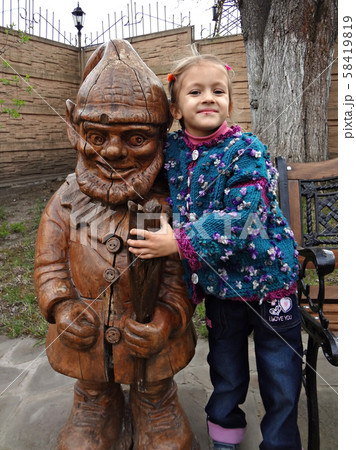 Little happy girl in a green knitted sweater stands next to a wooden gnome 58419819