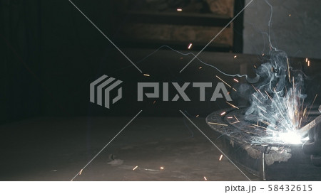 Forge workshop. Smithy. Worker in a welding hood helmet welds a part by electric welding. Sparks are 58432615