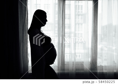 Silhouette of a pregnant woman holding her belly looking out a window with curtains Silhouette of a pregnant woman holding her belly looking out a window with curtains 58450225