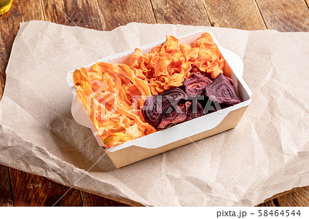 Paper box with homemade healthy vegetable chips from carrot and beetroot on wrapping paper on old wooden table. Natural healthy vegetable snack. Alternative to ready-made chips. 58464544
