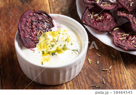 Close-up healthy homemade beetroot chips in white ceramic cup with sour cream sauce with herbs and on white plate on wooden background. Natural healthy vegetable snack. Alternative to ready-made chips 58464545