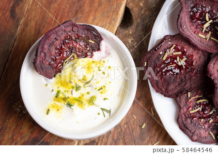 Close-up homemade beetroot chips in white cup with sour cream sauce with herbs and on white plate on wooden background. Natural healthy vegetable snack. Alternative to ready-made chips. Top view. 58464546