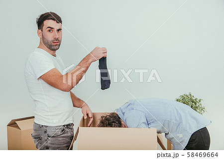 Wife and husband are busy, unpacking the carton boxes. Man is standing. holding black sock, looking 58469664