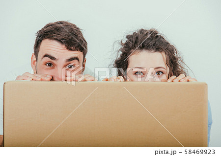 Funny wife and husband hiding behind the big carton box, together, looking over box at the camera. 58469923