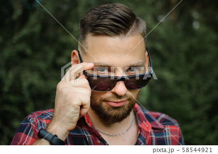 Outdoors portrait of young bearded man with sunglasses looking at the camera in the park on trees 58544921