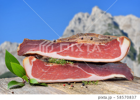 Typical South Tyrolean bacon divided into two halves lying on a rustic table in front of the alps 58570837