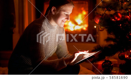 Portrait of smiling young woman sitting next to Christmas tree and typing message on tablet computer Portrait of smiling young woman sitting next to Christmas tree and typing message on tablet computer 58581690