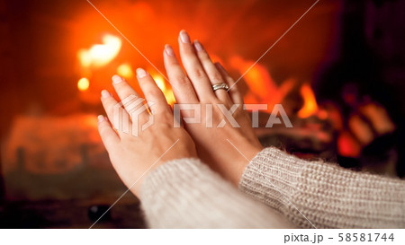 Closeup image of young woman in sweater warming her hands at burning fireplace at house 58581744