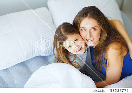 Beautiful mom and daughter are hugging, looking at camera and smiling while sitting on bed at home Beautiful mom and daughter are hugging, looking at camera and smiling while sitting on bed at home 58587775
