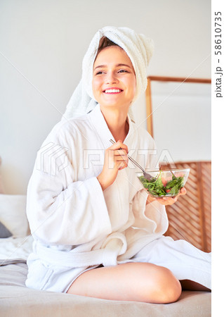 Picture of smiling woman in bathrobe with salad in bowl in her hands sitting on bed. Picture of smiling woman in bathrobe with salad in bowl in her hands sitting on bed. 58610735