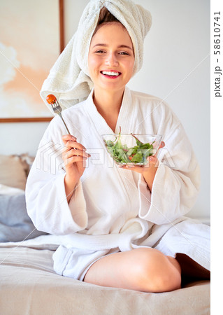 Image of young girl in bathrobe with salad in bowl in her hands sitting on bed. Image of young girl in bathrobe with salad in bowl in her hands sitting on bed. 58610741