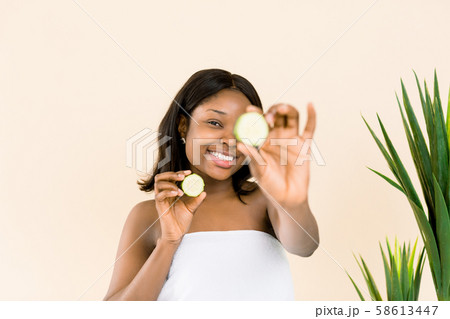Fun portrait of beautiful black african model with smooth skin holding a cucumber slice to her eye Fun portrait of beautiful black african model with smooth skin holding a cucumber slice to her eye 58613447
