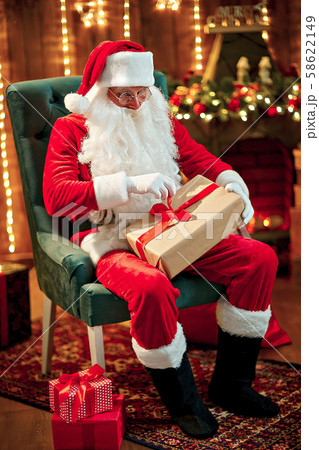 Photo of happy Santa Claus in eyeglasses sitting on chair, hold gift box Photo of happy Santa Claus in eyeglasses sitting on chair, hold gift box 58622149
