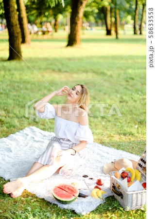 Young blonde woman having picnic on plaid and sitting in park with fruits. 58633558