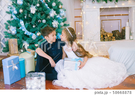 Children, a boy and a girl in a white ball gown near a Christmas tree in holiday dresses with gifts and silver confetti. Christmas mood Children, a boy and a girl in a white ball gown near a Christmas tree in holiday dresses with gifts and silver confetti. Christmas mood 58668878