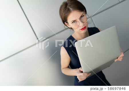 businesswoman holds laptop in her hands, looks in disbelief at the camera, portrait against a gray businesswoman holds laptop in her hands, looks in disbelief at the camera, portrait against a gray 58697846