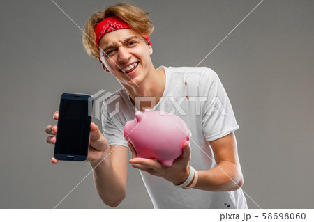 teenager with piggy bank posing against gray, young man in a light t-shirt against a gray wall 58698060