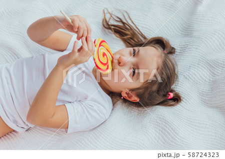 Gorgeous kid with funny pigtails is licking red-yellow candy on stick, smiling lovely to the camera. Gorgeous kid with funny pigtails is licking red-yellow candy on stick, smiling lovely to the camera. 58724323