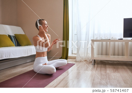 Cheerful caucasian young short-haired woman sit on mat at home and listen to music with headphones 58741407