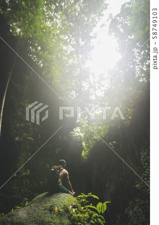 Man wearing backpack sitting on rock under trees in Bali, Indonesia Man wearing backpack sitting on rock under trees in Bali, Indonesia 58749103